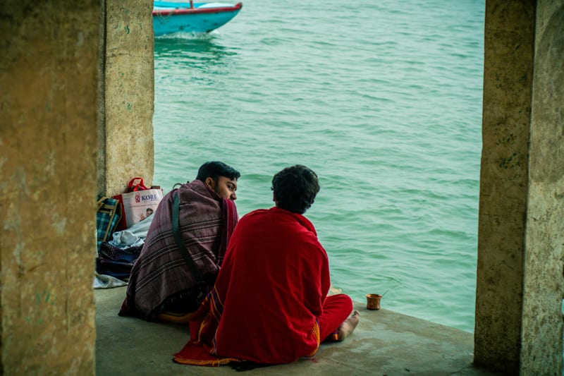 Two figures in maroon and red shawls seated by the Ganges, framed by stone columns